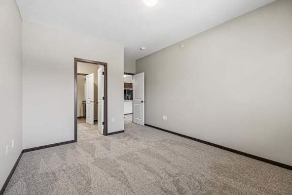 Carpeted apartment bedroom with gray walls, dark trim, open doors, and a view toward the kitchen.