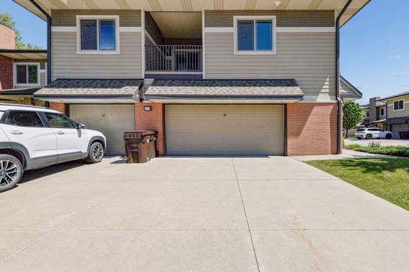 Apartment exterior with attached garages, concrete driveway, upper balcony, tan siding, and brick accents.