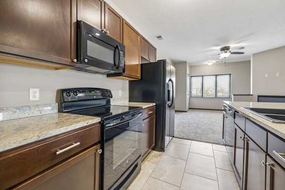 Apartment kitchen with dark wood cabinets, granite-style countertops, black appliances, tile flooring, and open living room view.