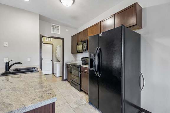 Apartment kitchen with granite-style counters, dark cabinets, black appliances, tile flooring, and laundry room access.