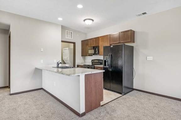 Open apartment kitchen with granite-style island, dark cabinets, black appliances, tile flooring, and carpeted living area.