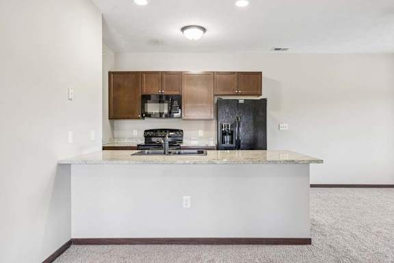 Open apartment kitchen with granite-style breakfast bar, dark cabinets, black appliances, and carpeted living area.