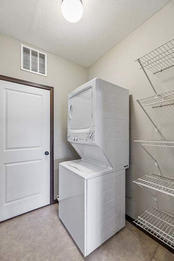 Apartment laundry room with stacked washer and dryer, wire shelving, overhead light, and neutral walls.