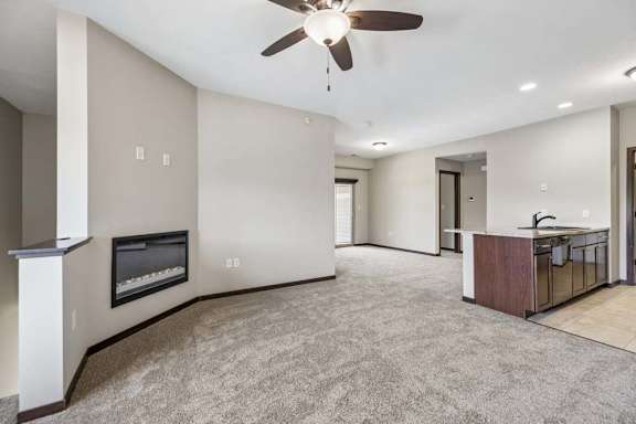 Open apartment living area with carpet, ceiling fan, wall fireplace, and kitchen with dark cabinets and island.
