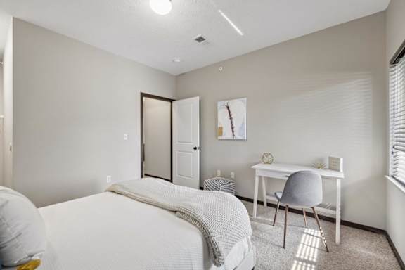 Staged apartment bedroom with carpet, bed, desk, window with blinds, and neutral walls.