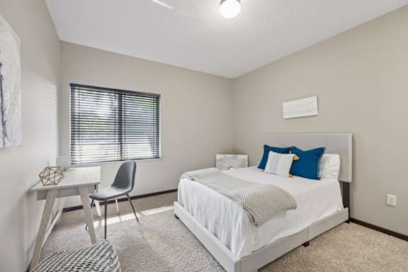 Bright staged apartment bedroom with carpet, upholstered bed, desk, window with blinds, and neutral finishes.