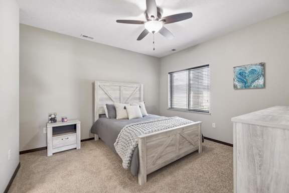 Staged apartment bedroom with ceiling fan, carpet, wood-look bed frame, dresser, and window with blinds.