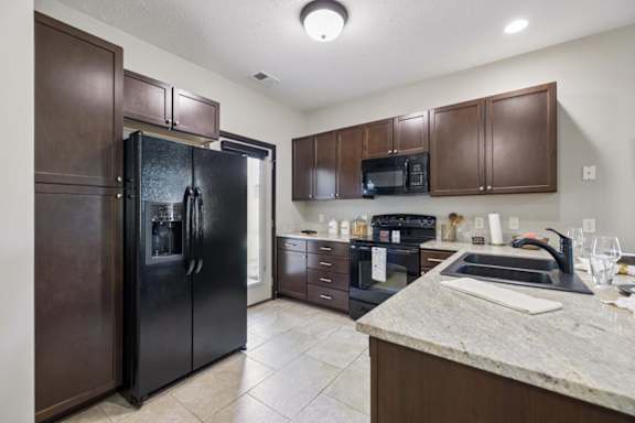 Apartment kitchen with dark wood cabinets, speckled counters, black appliances, tile floor, and windowed door.
