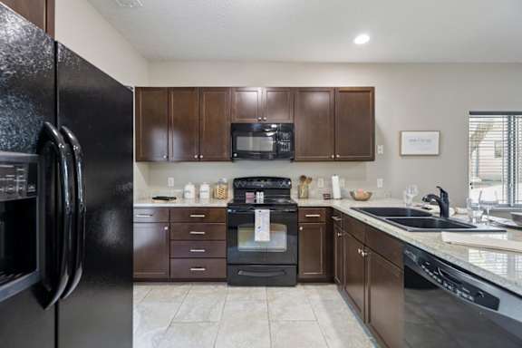 Staged apartment kitchen with dark wood cabinets, black appliances, speckled counters, and tile flooring.