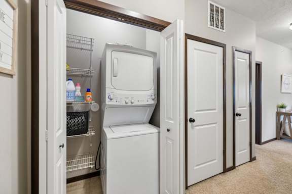 Laundry closet with stacked washer and dryer, wire shelving, cleaning storage, and carpeted hallway.