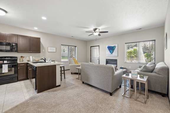 Staged open living room and kitchen with carpet, ceiling fan, fireplace, gray seating, and breakfast bar.