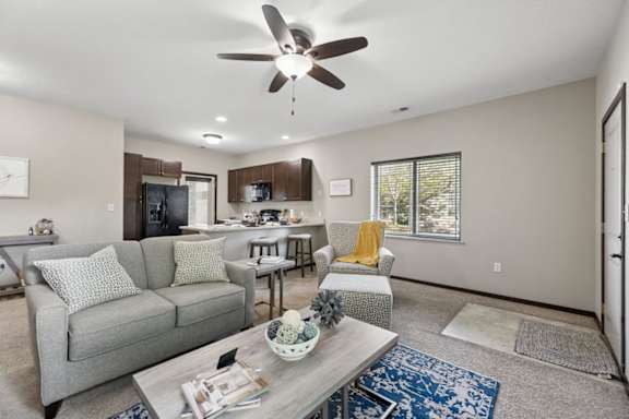 Staged apartment living room with sofa, armchair, ceiling fan, kitchen view, and entry door with tile landing.