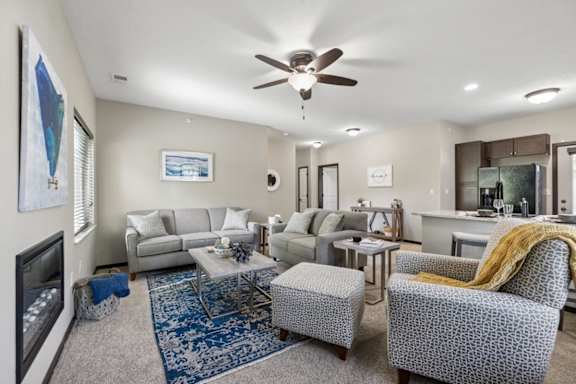 Staged apartment living room with sofa, armchair, ceiling fan, kitchen view, and entry door with tile landing.