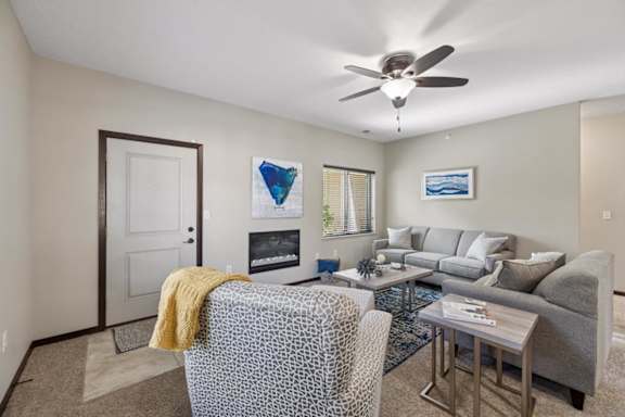 Staged apartment living room with sofa, armchair, ceiling fan, kitchen view, and entry door with tile landing.
