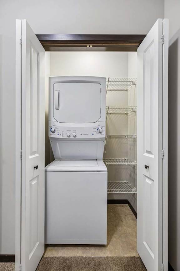 Apartment laundry closet with stacked washer and dryer, wire shelving, and double doors.