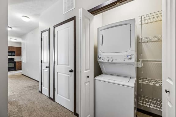 Hallway laundry closet with stacked washer and dryer, wire shelving, carpeted hall, and nearby kitchen.
