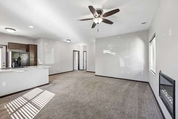 Open apartment living area with carpet, ceiling fan, fireplace, windows, and kitchen with breakfast bar.
