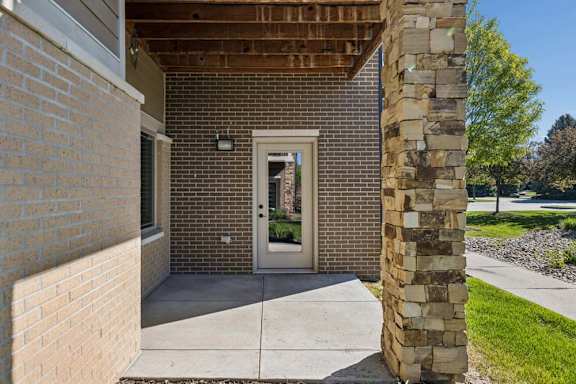 Covered apartment patio entry with brick walls, stone column, concrete walkway, grass, and landscaped grounds.