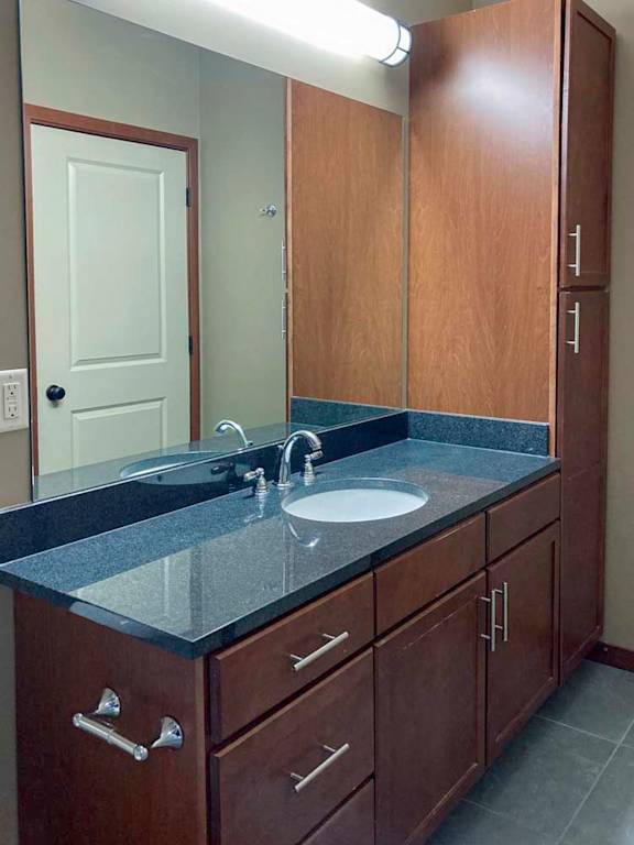 A bathroom with a wide granite vanity sink, tall storage cabinet, and large mirror.