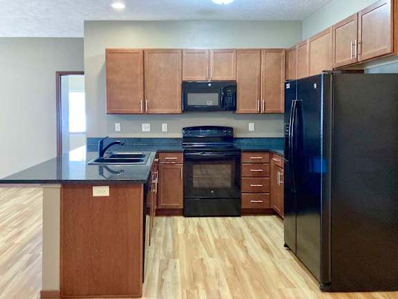 A modern kitchen with brown cabinets, dark granite counters, and black appliances.
