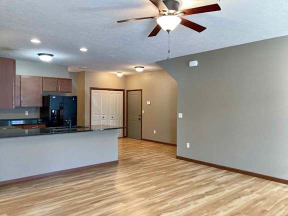 A living room with hardwood-style flooring and a ceiling fan, open to a modern kitchen with brown cabinets, dark granite counters, and black appliances.
