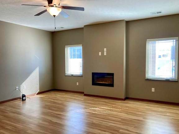 A living room with hardwood-style flooring, a ceiling fan, large windows, and an electric fireplace.
