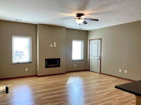 A living room with hardwood-style flooring, a ceiling fan, large windows, and an electric fireplace.