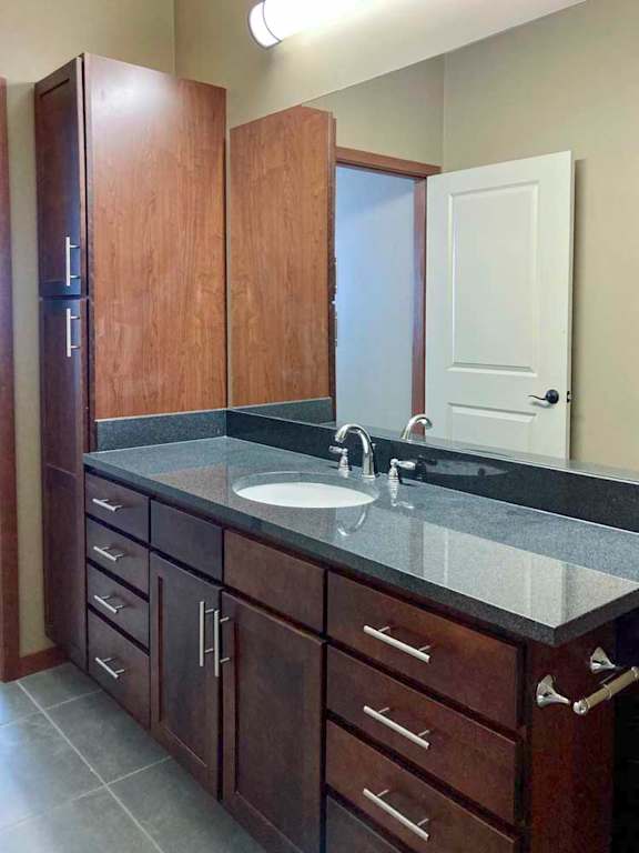 A long bathroom with a wide granite vanity sink, oversized mirror, and tall storage cabinet.