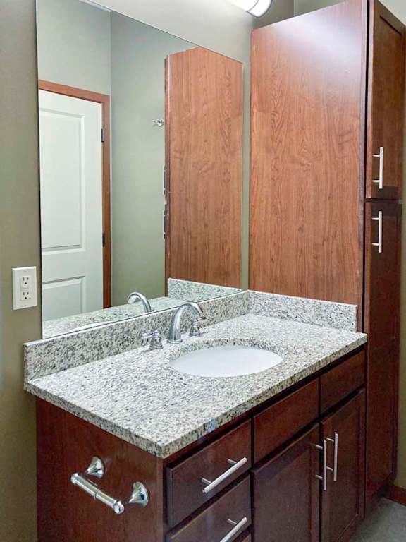 A bathroom with a granite vanity sink, a tall storage cabinet, and an oversized mirror with overhead lighting.