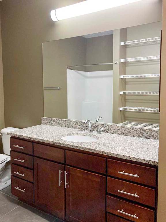 A bathroom with a wide granite vanity sink, dark cabinets, an oversized mirror with overhead lighting, and a walk-in shower.