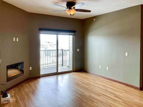 A living room with hardwood-style flooring, a sliding glass door to the balcony, a ceiling fan, and an electric fireplace.