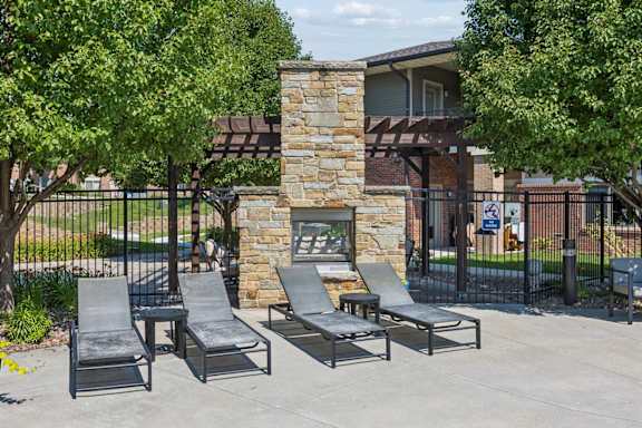 Poolside lounge area at Villas of Omaha at Butler Ridge with chaise seating, a stone fireplace, pergola, and mature trees.