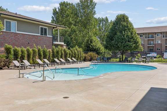 Outdoor swimming pool at Villas of Omaha at Butler Ridge with lounge chairs, entry steps, mature trees, and apartment buildings.