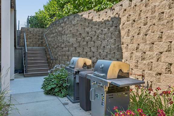 Outdoor grilling amenity at Villas of Omaha at Butler Ridge with two grills, landscaped greenery, and nearby stairs.