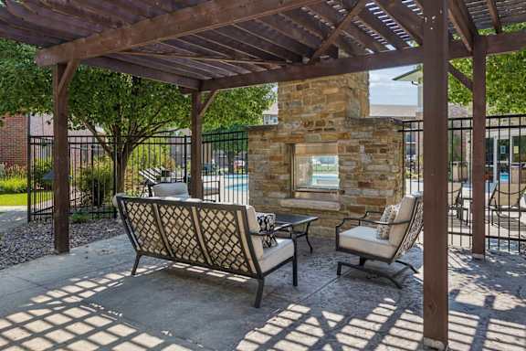 Pergola-covered outdoor lounge at Villas of Omaha at Butler Ridge with cushioned seating and a stone fireplace beside the pool.