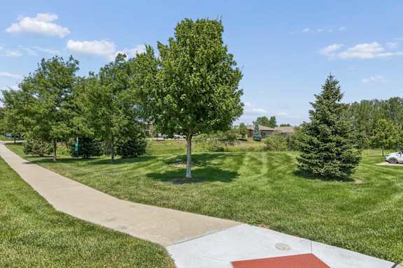 Landscaped walking path at Villas of Omaha at Butler Ridge winding through green lawn and mature trees near apartment buildings.