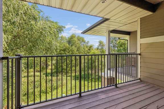 Covered balcony with wood-look decking, black metal railing, and a peaceful view of mature green trees.
