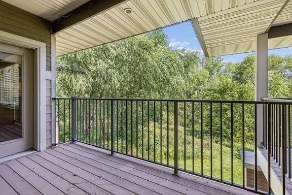 Covered balcony with wood-look decking, black metal railing, and a peaceful view of mature green trees.