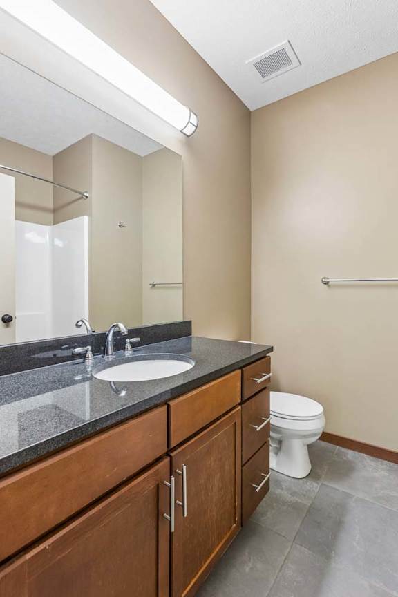 Bathroom with wood vanity, dark countertop, large mirror, bright vanity light, neutral walls, and gray tile flooring.