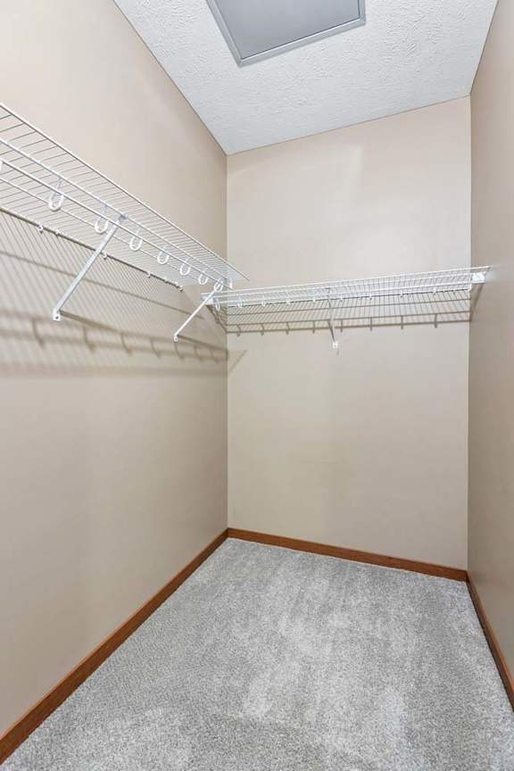 Spacious apartment walk-in closet with beige walls, carpeted flooring, wire shelving, and overhead lighting.