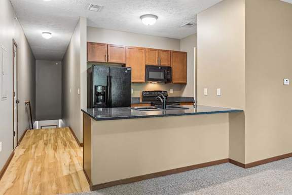 Open kitchen with wood cabinetry, black appliances, breakfast bar, dark countertops, and adjacent wood-look flooring.