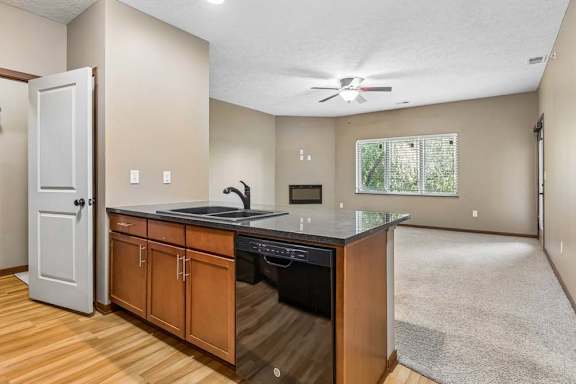 Open kitchen and living area with wood cabinetry, dark countertops, double sink, ceiling fan, fireplace, and large window.