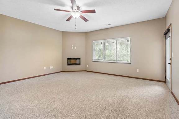 Bright living room with neutral carpet, ceiling fan, large window with blinds, and a fireplace.