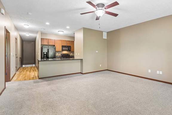Open living area with neutral carpet, ceiling fan, breakfast bar, wood cabinetry, and black kitchen appliances.