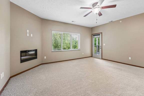 Bright living room with neutral carpet, ceiling fan, fireplace, large window with blinds, and balcony access.