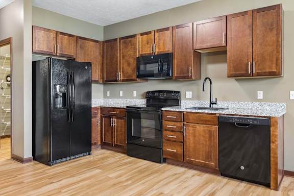 Apartment kitchen with warm wood cabinetry, speckled countertops, black appliances, wood-look flooring, and a tall curved faucet.