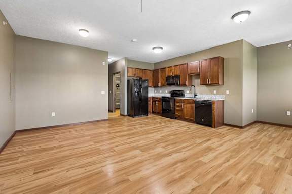 Spacious open apartment living and kitchen area with wood-look flooring, neutral walls, wood cabinetry, and black appliances.