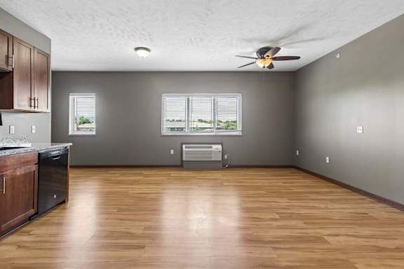 Empty apartment living area with wood-look flooring, neutral walls, ceiling fan, large windows, and an open kitchen.