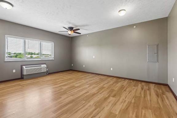 Open apartment living area with wood-look flooring, neutral walls, ceiling fan, wide windows, and a wall-mounted air conditioner.