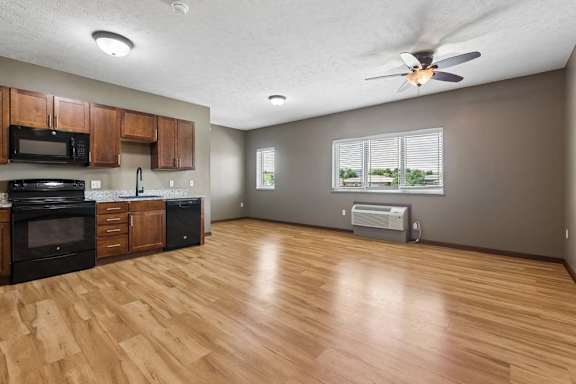 Open-concept apartment kitchen and living area with wood-look flooring, dark appliances, wood cabinetry, ceiling fan, and large windows.